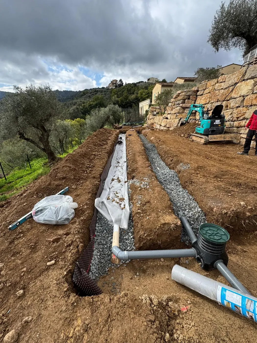 Canalisations et regard dans les tranchées gravillonnées, avec une mini-pelle près du mur en pierre à Châteauneuf-de-Grasse